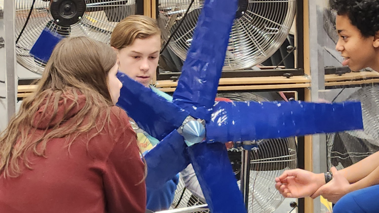 Three students crouch around windmill device covered in blue tape